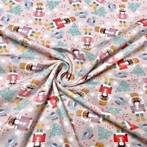 May include: Pink fabric with a repeating pattern of nutcracker figures, candy canes, and Christmas trees. The nutcrackers are wearing different colored hats and coats.