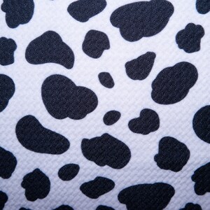 May include: Close-up of a white fabric with a repeating pattern of black, irregular shapes resembling cow spots. The fabric has a textured surface, and the black spots vary in size and shape, creating a playful, animal-inspired design.