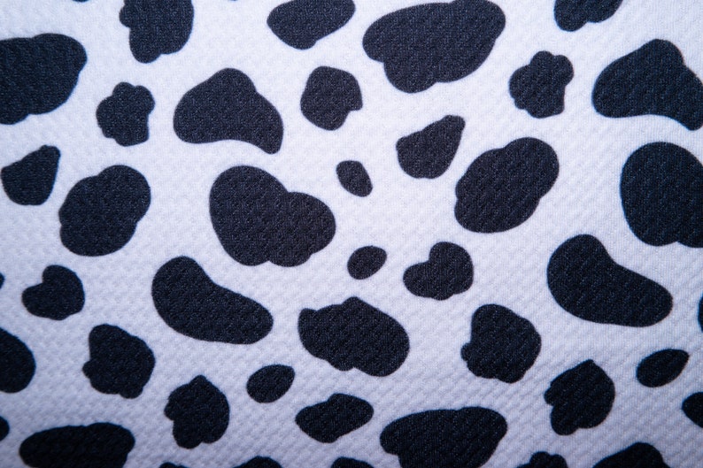 May include: Close-up of a white fabric with a repeating pattern of black, irregular shapes resembling cow spots. The fabric has a textured surface, and the black spots vary in size and shape, creating a playful, animal-inspired design.