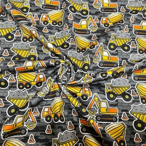 May include: Fabric with a repeating pattern of construction vehicles, including dump trucks, excavators, and cement mixers. The vehicles are yellow and orange with black details, set against a gray brick-like background. White outlines define each vehicle.