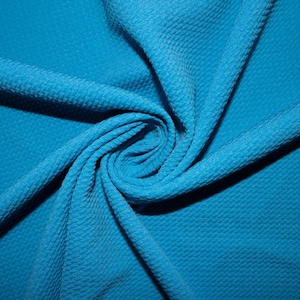 Puede incluir: Primer plano de una tela de color turquesa brillante con un patrón texturizado de tejido de gofre. La tela está doblada y retorcida, creando un efecto de remolino.