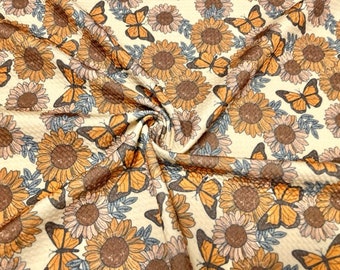 May include: Cream-colored fabric with a repeating pattern of sunflowers and butterflies. The sunflowers are in shades of brown and yellow, with orange butterflies scattered throughout. The fabric has a textured appearance.