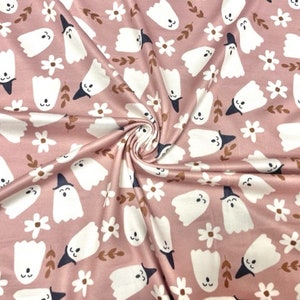 May include: Pink fabric with a repeating pattern of white ghosts wearing black witch hats, white daisies, and brown leaves. The fabric is folded, creating shadows and texture. Ideal for Halloween or spooky-themed crafts.