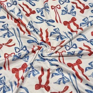 Puede incluir: Tela blanca con un patrón repetido de lazos y cintas rojos y azules. Los lazos son de estilo acuarela, con un aspecto suave y texturizado. La tela está doblada, creando sombras y profundidad.