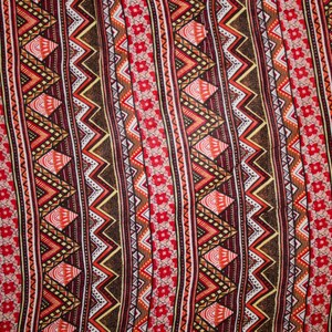 May include: Close-up of a fabric with a repeating geometric pattern. The design features alternating vertical stripes of zigzags, diamonds, and floral elements in shades of red, brown, orange, and white. The overall aesthetic is reminiscent of tribal or ethnic textiles.