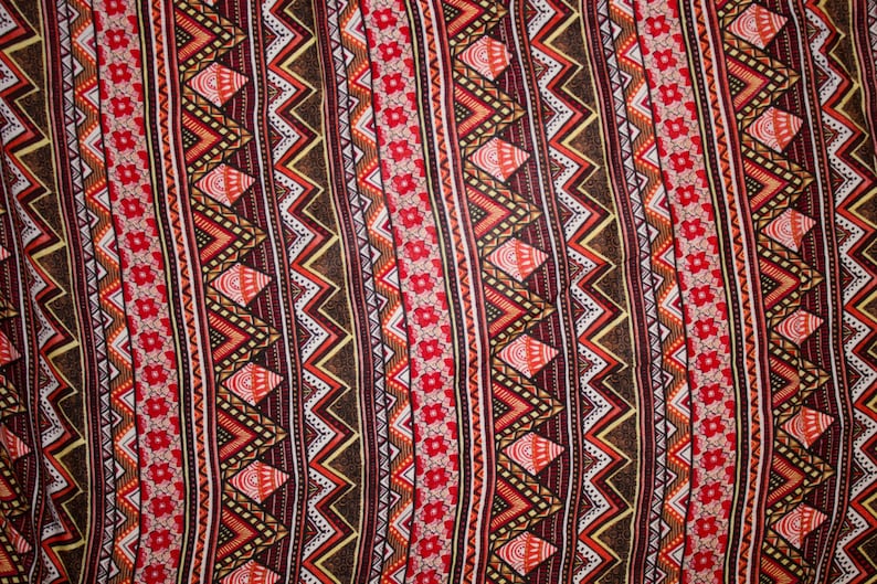 May include: Close-up of a fabric with a repeating geometric pattern. The design features alternating vertical stripes of zigzags, diamonds, and floral elements in shades of red, brown, orange, and white. The overall aesthetic is reminiscent of tribal or ethnic textiles.