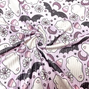 May include: A light purple fabric with a repeating Halloween-themed pattern. The design includes smiling ghosts, bats, candy corn, crescent moons, flowers, and stars. The fabric has a ribbed texture.