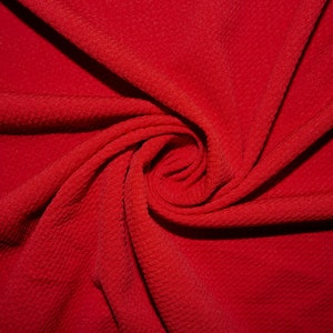 Puede incluir: Primer plano de una tela roja vibrante, dispuesta en espiral. El material tiene una superficie texturizada, con un patrón sutil visible. Los pliegues y curvas de la tela crean una composición dinámica y visualmente atractiva. Ideal para costura.