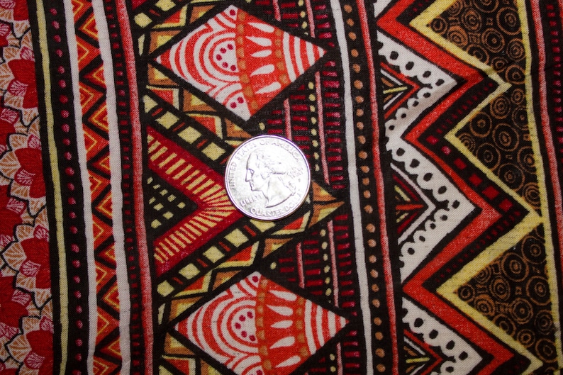 May include: Close-up of a fabric with a vibrant geometric pattern. The design features repeating shapes in red, orange, yellow, and black, with a quarter coin for scale. The fabric has a tribal or ethnic aesthetic.