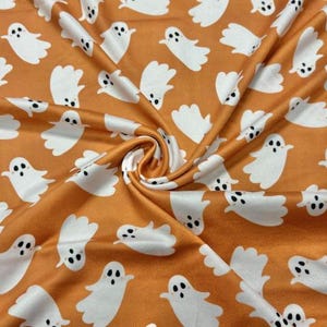 May include: A fabric featuring a repeating pattern of white ghosts with black eyes and mouths scattered across a solid orange background. The material is gathered in the center, creating soft folds and shadows that highlight the ghost print.