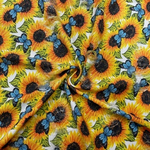 May include: Fabric with a repeating pattern of sunflowers and blue butterflies on a white background. The sunflowers are yellow with dark brown centers, and the butterflies have blue wings. The fabric is folded, showing the texture and design.