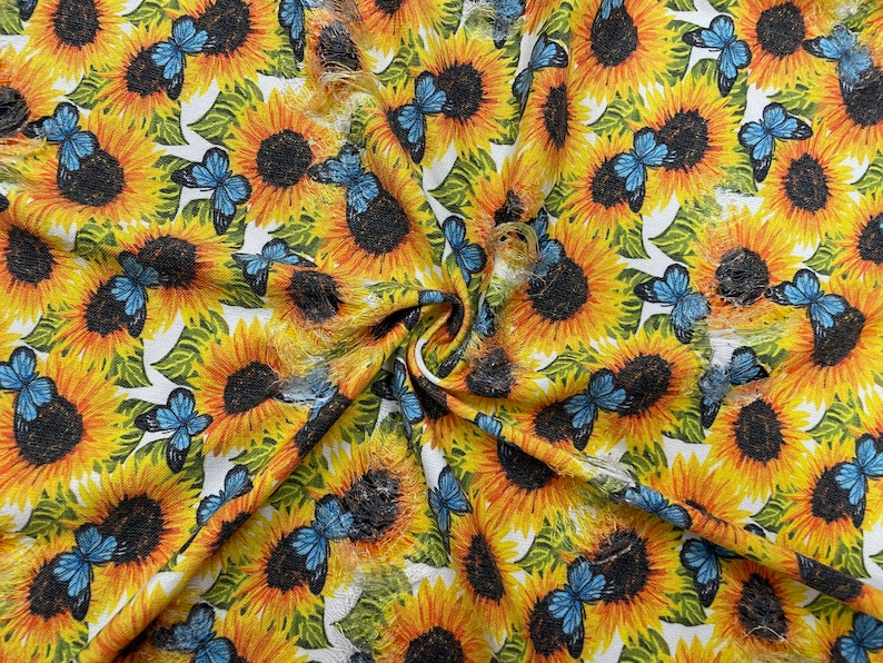 May include: Fabric with a repeating pattern of sunflowers and blue butterflies on a white background. The sunflowers are yellow with dark brown centers, and the butterflies have blue wings. The fabric is folded, showing the texture and design.