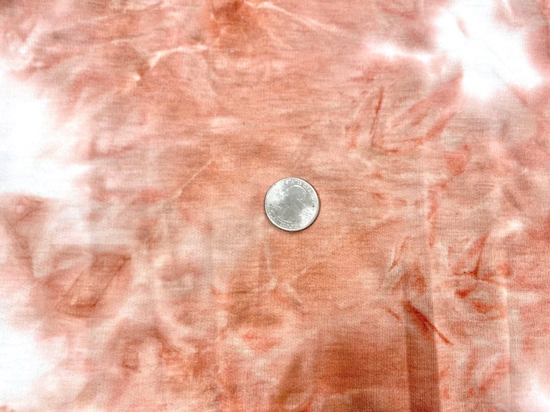 May include: A close-up of a tie-dye fabric in shades of white and orange. A US quarter is placed on the fabric to show the scale.