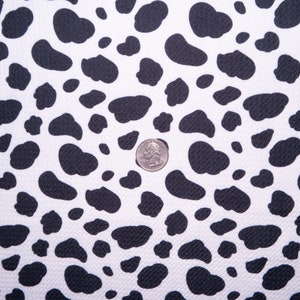 May include: White fabric with a repeating pattern of black, irregular shapes, resembling a cowhide design. A US quarter coin is placed on the fabric for scale. The fabric has a textured surface.