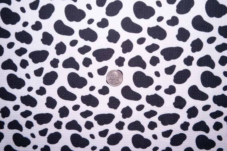 May include: White fabric with a repeating pattern of black, irregular shapes, resembling a cowhide design. A US quarter coin is placed on the fabric for scale. The fabric has a textured surface.