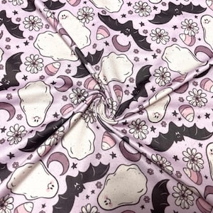 May include: A light purple fabric with a repeating pattern of Halloween-themed illustrations. The design includes white ghosts, black bats, pink candy corn, crescent moons, and daisies. The fabric is folded, creating a textured appearance.