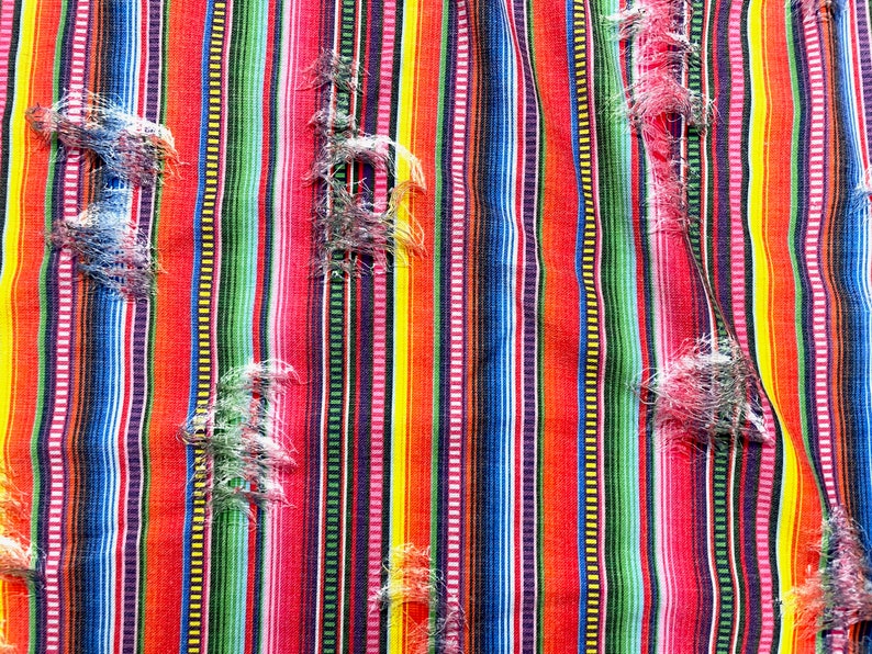 K&ouml;nnte beinhalten: Nahaufnahme eines farbenfrohen Stoffes mit Streifen und einem Used-Look-Design. Der Stoff weist vertikale Streifen in Rot, Orange, Gelb, Gr&uuml;n, Blau und Lila auf. Der Stoff hat ausgefranste Bereiche, die Textur und visuelles Interesse hinzuf&uuml;gen.