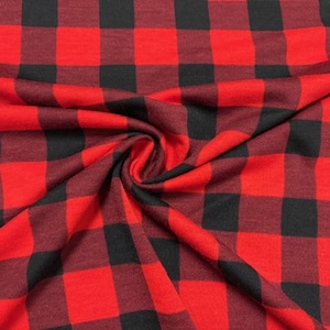 May include: Close-up of a red and black buffalo plaid fabric. The fabric is folded and draped, showing the pattern's repeating large squares. The colors are a vibrant red and a deep black, creating a classic, rustic look. The material appears soft.
