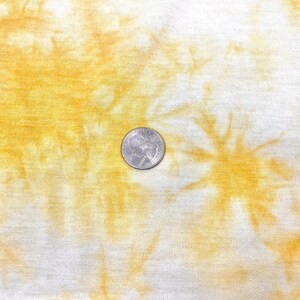 May include: A close-up of a white fabric with a yellow tie-dye pattern. A US quarter is placed on the fabric for scale.