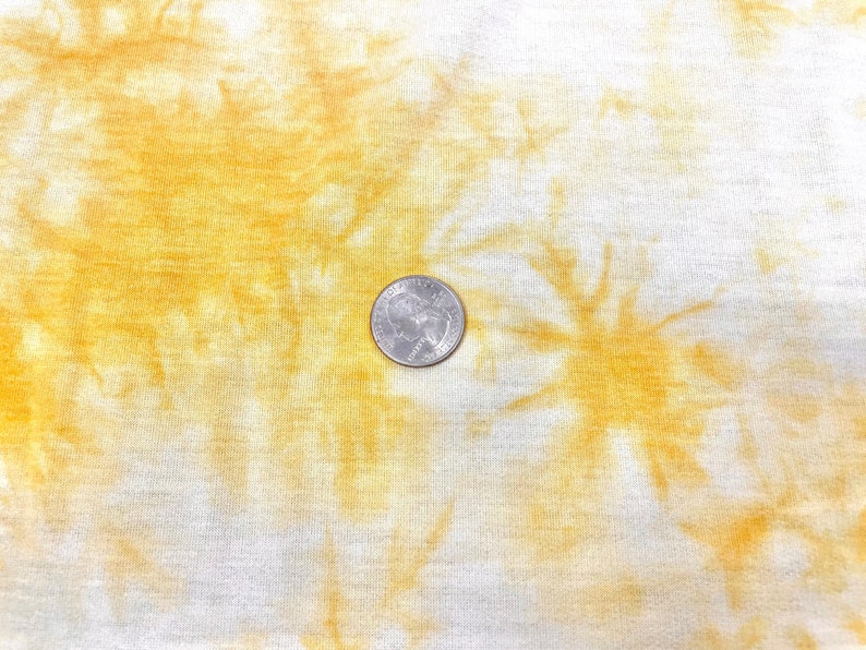 May include: A close-up of a white fabric with a yellow tie-dye pattern. A US quarter is placed on the fabric for scale.