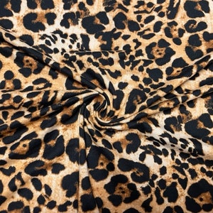 May include: Close-up of a fabric with a leopard print pattern. The fabric features a tan background with black spots of varying sizes and shapes. The fabric is folded and draped, showing the texture and flow of the material.