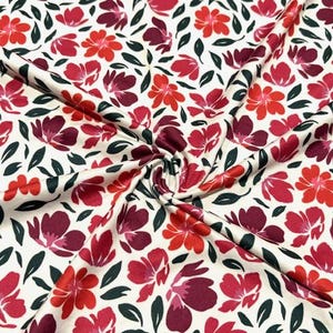 May include: Cream-colored fabric with a repeating floral pattern. The flowers are in shades of red and burgundy, with dark green leaves. The fabric is gathered in the center, creating folds and shadows.