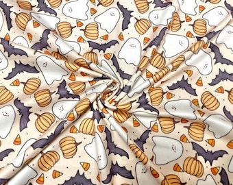 May include: A cream-coloured fabric with a repeating pattern of orange pumpkins, white ghosts, and purple bats. The pattern is set against a light beige background.