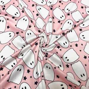 May include: Pink fabric with a repeating pattern of white cartoon ghosts and black stars. The ghosts have two black dot eyes. The fabric is folded and draped, showing the texture and sheen of the material.