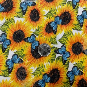 May include: Fabric with a repeating pattern of yellow sunflowers and blue butterflies. The sunflowers have dark brown centers and green leaves. A U.S. quarter coin is visible on the fabric, providing a size reference.