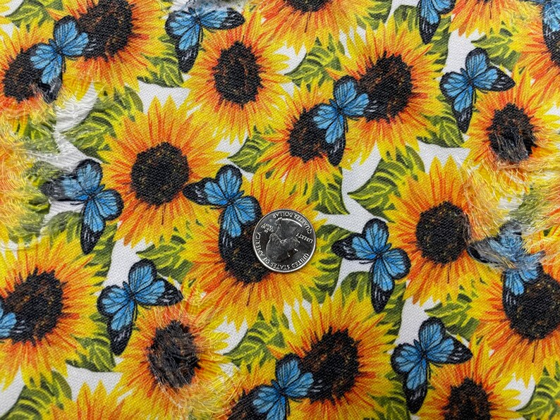 May include: Fabric with a repeating pattern of yellow sunflowers and blue butterflies. The sunflowers have dark brown centers and green leaves. A U.S. quarter coin is visible on the fabric, providing a size reference.