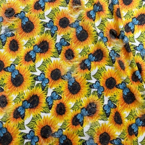 May include: A fabric with a repeating pattern of sunflowers and blue butterflies. The sunflowers are yellow with dark brown centers, and the butterflies have blue wings. The background is white with green leaves.