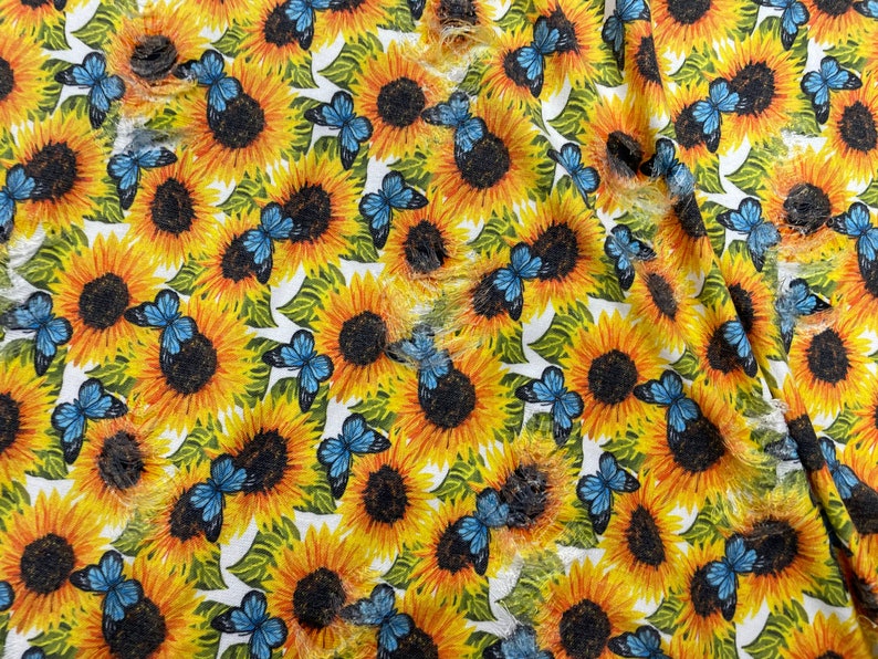 May include: A fabric with a repeating pattern of sunflowers and blue butterflies. The sunflowers are yellow with dark brown centers, and the butterflies have blue wings. The background is white with green leaves.
