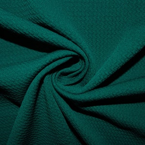 Op de afbeelding: Een close-up van een diep teal stof met een textuur, een wafelachtig patroon. De stof is gevouwen en gedrapeerd, waardoor een zachte en uitnodigende uitstraling ontstaat.
