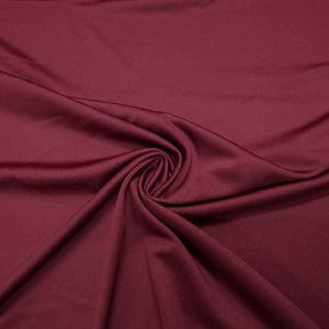 May include: Close-up of a deep burgundy fabric with a smooth, flowing texture. The material is gathered and swirled, creating soft folds and shadows. The rich color and drape suggest a luxurious textile suitable for apparel or home decor.