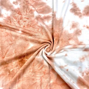 May include: A close-up of a soft, peach-colored fabric with a tie-dye pattern. The fabric is folded and twisted, showing off the swirling pattern.