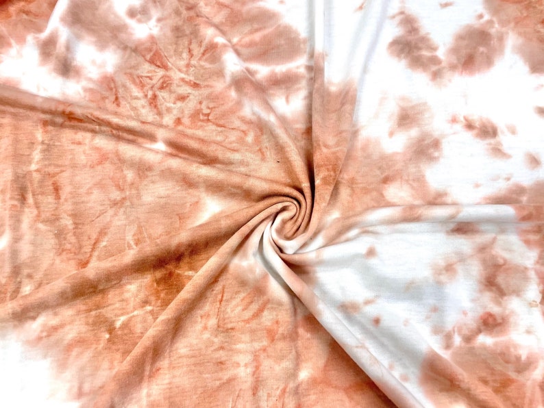 May include: A close-up of a soft, peach-colored fabric with a tie-dye pattern. The fabric is folded and twisted, showing off the swirling pattern.