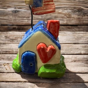 May include: A small ceramic house figurine with a cream-colored facade, a blue roof, and a red chimney. A red heart is attached to the front, and a blue door is visible. A miniature American flag on a wire pole is attached to the roof.