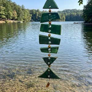 May include: A hanging green ceramic wind chime with a series of geometric shapes, including triangles and rectangles, strung together with wooden beads. The chime is suspended in front of a lake with a clear blue sky.