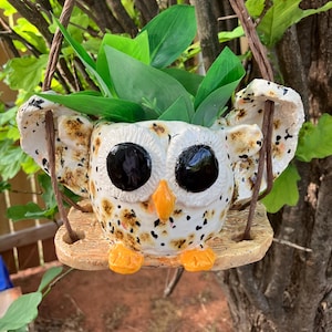 May include: A ceramic owl planter with black eyes, an orange beak, and feet. The owl is white with brown speckles and wings. It hangs from a brown rope and holds green leaves.
