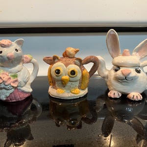 May include: Three ceramic teapots in the shape of animals. A cat teapot with a pink dress and flowers, an owl teapot with large yellow eyes, and a bunny teapot with long ears and pink paws.