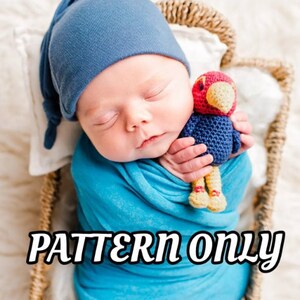 Puede incluir: Un bebé durmiendo con un gorro azul anudado y una manta azul, sosteniendo un juguete de pájaro de crochet rojo y azul. El bebé está acostado en una cesta tejida.