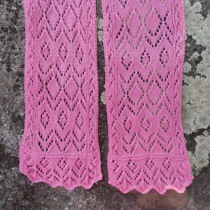 May include: Two pink crocheted scarves with a lacy diamond pattern. The scarves are laid out flat on a textured gray surface.