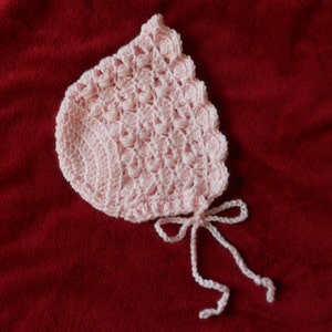 May include: A pink crocheted baby bonnet with a scalloped edge and a tied bow.