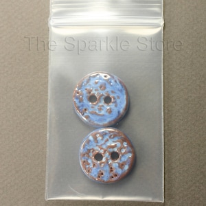 May include: Two blue ceramic buttons with brown speckles and two holes each. The buttons are in a clear plastic bag.