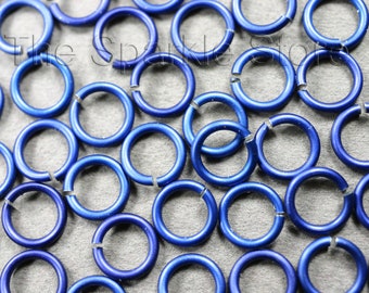 May include: A collection of small, open, blue jump rings scattered across a gray surface. The rings are metallic and have a slight sheen. The image is a close-up, showing the details of the rings.