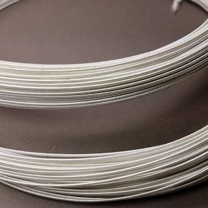 May include: Two coils of silver-colored wire, tightly wound, are displayed against a dark background. The wire appears to be smooth and metallic, with a slight sheen. One coil is partially visible, while the other is in the foreground, showing the full circular form. A blue thread secures one coil.