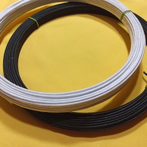 May include: Two coiled elastic cords, one white and one black, are displayed against a yellow background. The cords are bound with green bands. The image is well-lit, highlighting the texture and color of the materials.