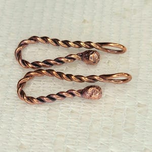 May include: Two copper-colored, twisted metal hair accessories. Each piece has a U-shape with a small, textured ball at the center and a loop at the end. The accessories are displayed on a white, textured surface.