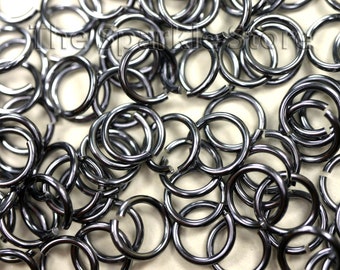 May include: Close-up of a pile of open jump rings. The rings are a dark, metallic gray color and are open, ready to be used in jewelry making or other crafts. The rings are scattered across the surface.