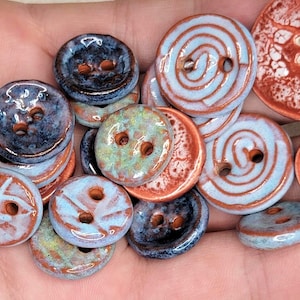 May include: A selection of handmade ceramic buttons in various sizes and designs. The buttons feature a mix of colours, including blue, brown, and green, with spiral and textured patterns. Each button has two or four holes for sewing.